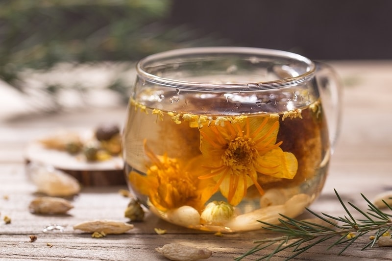 Glass of herbal tea containing a suspended flower. Bits of herbs and flowers surround the glass.